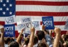 People holding up signs saying "Vote" in front of a US flag